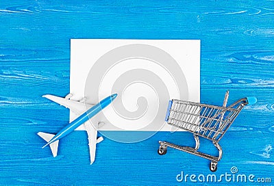 Model of airplane, grocery cart and blank sheet of paper on the blue wooden background. Travel concept. Ticket purchase. Stock Photo