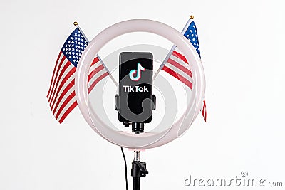 Tiktok logo on a smartphone attached to a round lamp with a tripod and two American flags on a white background. Donald Trump to Editorial Stock Photo