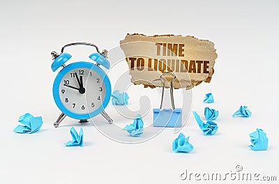 On a white surface, an alarm clock, an office clip, crumpled paper and a cardboard sign with the text -Time to Liquidate Stock Photo