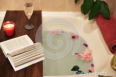 Bath with flower petals. Book, candles and glass of wine on a wood tray. Organic Spa Relaxation in comfort cozy bathroom Stock Photo