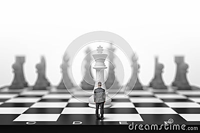 A tiny businessman stands on a giant chessboard in front of a white king in a back view. Stock Photo