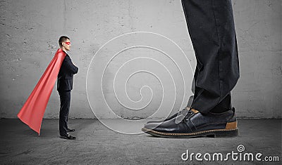 A tiny businessman in a superhero cape stands facing giant man with only his feet seen. Stock Photo