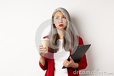 Tired asian female office worker holding clipboard and paper cup, drinking coffee and exhaling with exhausted face Stock Photo