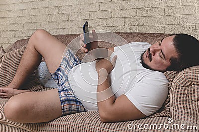 A tired and bored slacker in boxer shorts is addicted on the phone during a lazy day lying on the couch Stock Photo