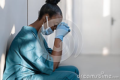 Tired depressed female african nurse wearing face mask sits on hospital floor. Stock Photo