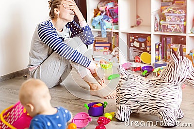 Tired of everyday household mother sitting on floor with hands on face. Kid playing in messy room. Scaterred toys and disorder. Stock Photo