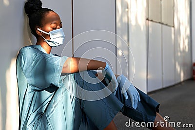 Tired exhausted woman african nurse wear face mask gloves sit on hospital floor. Stock Photo