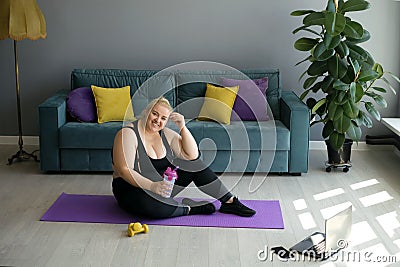 A tired happy fat woman is sitting on a training mat holding a bottle of water in her hand. Stock Photo