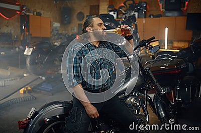 Tired man biker drinking beer from bottle after hard repair work in garage Stock Photo
