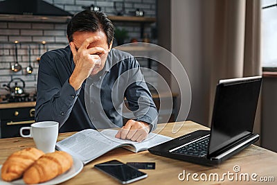 Tired man sit at table in kitchen. He cover face with hand. Man work. He has headache. Stock Photo