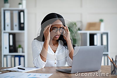 Tired millennial black businesswoman suffering from terrible headache at desk in office Stock Photo