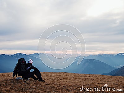 Tired out tourist in black with backpack is sitting on stone in meadow and watching into misty valley. Autumn in mountains. Stock Photo