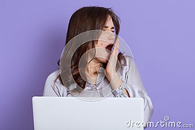 Tired sleepy woman yawning and covering mouth with hand, working on laptop sitting against lilac background, wearing elegant Stock Photo