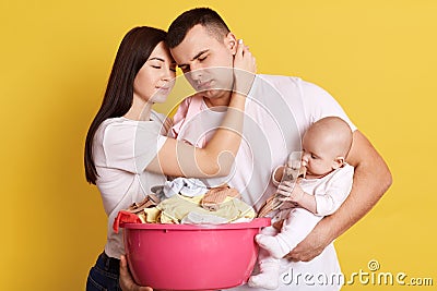 Tired troublesome parents crying newborn baby and basin full of clean clothing, have much domestic work, posing isolated on yellow Stock Photo
