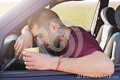 Tired unshaven male leans on wheel and holds paper cup of coffee, has sleepy expression, sits in car, slumbers in automobile, bein Stock Photo