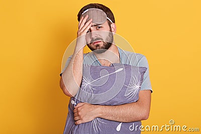 Tired unshaven man standing against yellow background, holding grey pillow, touching his head, wearing gray t shirt and blindfold Stock Photo