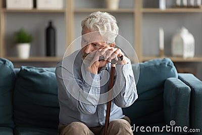 Tired upset mature man holding walking stick, sitting on couch Stock Photo