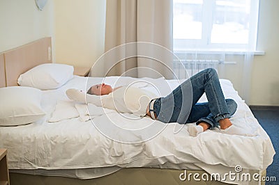 Tired woman is lying in bed with her arm on head and eyes. Young woman with long hair, wears jeans Stock Photo