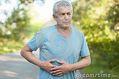 Tired wrinkled senior male keeps hands on body, feels pain, catches breath after long jogging, dressed in casual t shirt, stands o Stock Photo