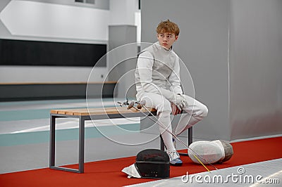 Tired young fencer in special costume sitting on bench and looking aside Stock Photo