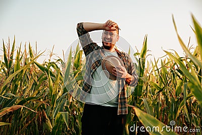 Tired young man stand in cornfield, took off hat, wipe sweat from forehead, front view, looking away. Stock Photo