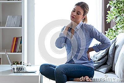 Tired young woman with shoulder and back pain sitting on the couch at home. Stock Photo