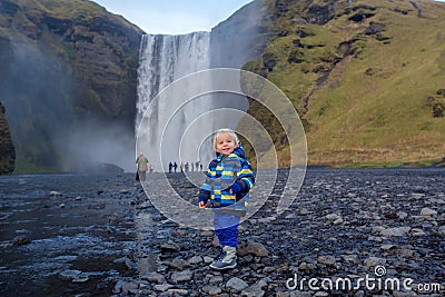 Toddler boy, playing with rocks on the river in fronf of Skogafoss waterfall in Iceland on a sunset cloudy day Stock Photo