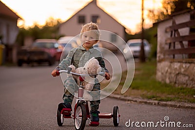 Toddler child, blond boy, riding tricycle in a village small road on sunset Stock Photo