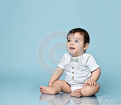 Little toddler in white bodysuit as a vest with bow-tie, barefoot. He is sitting on the floor against blue background. Close up Stock Photo