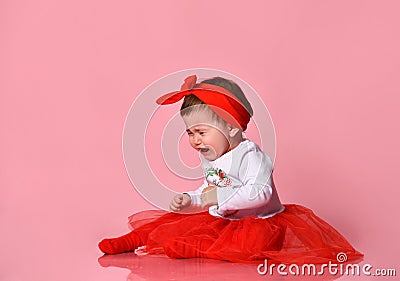Toddler in white bodysuit, red headband, poofy skirt. She crying, sitting on floor against pink studio background. Close up Stock Photo