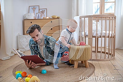 Father in a checkered shirt layng on the floor and reading a book, his little son playing next to him Stock Photo