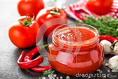 Tomato paste, puree in glass jar and fresh tomatos on dark background. Hot vegetable sauce with chili pepper ant tomatoes Stock Photo