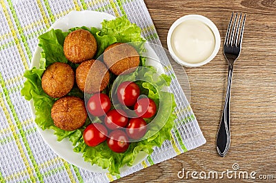 Tomatoes and round fried pies on lettuce in dish on napkin, bowl with mayonnaise and fork on wooden table. Top view Stock Photo