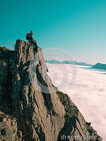 Toned image adult man with backpack sitting, legs dangling on the edge of a cliff and looks into the distance against the blue sky Stock Photo