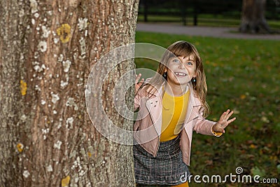 Toothless funny cute kid girl playing hide and seek in the autumn Park. Looks out from behind a tree with a playful smile and Stock Photo