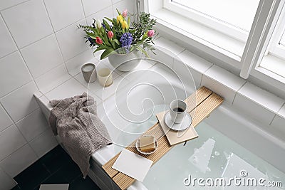 Top view of bathtub with foam. Book, coffee and soap on a tray. Fresh spring flowers with tulips, hyacinth and candles Stock Photo