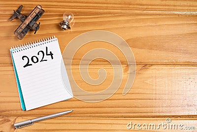 Top view of 2024 blank list on notebook on wooden table with miniature airplane, small light bulb and pen. New year goals and Stock Photo