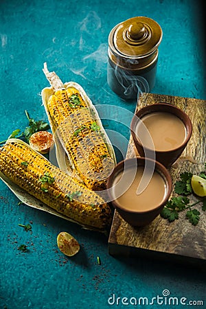 Steaming Hot Milk Tea served with roasted grilled corn on blue background. Two Delicious Grilled Corncobs and Two Cups of Chai. Stock Photo