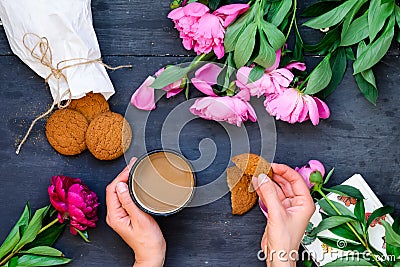 Top view close up female hands holding mug of coffee and cookies surrounded with peonies flowers. Coffee break concept. Selective Stock Photo