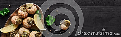 Top view of delicious cooked escargots with lemon and tweezers on black wooden table, panoramic shot. Stock Photo