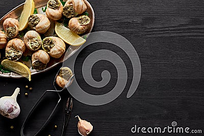 Top view of delicious cooked escargots with lemon and tweezers on black wooden table. Stock Photo