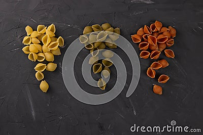 Top view of dry conchiglie pasta on black board background. Three triangular slides with pasta conchiglie spread out in colors, Stock Photo