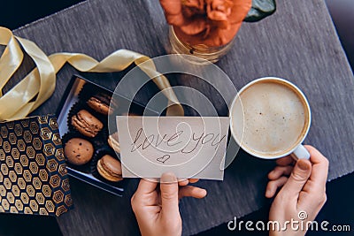 Top view female hands with coffee cup and greeting card with handwrite Love you words. Box with macaroons and flower on gray Stock Photo