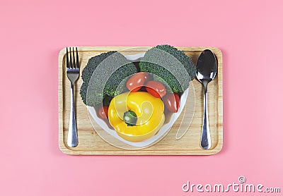 Flat lay of vegetables capsicum, broccoli and tomato in heart shape plate with fork and spoon in wooden tray on pink background Stock Photo