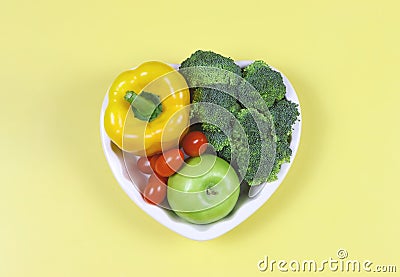 Flat lay of vegetables capsicum, broccoli and tomatoes and apple in heart shape plate on yellow background with copy space, Stock Photo