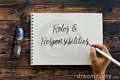Top view of glasses and hand holding pen writing Roles and Responsibilities on notebook on wooden background Stock Photo