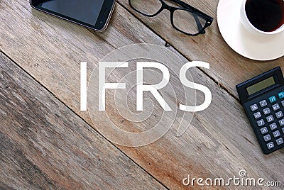 Top view of mobile phone, sunglasses, a cup of coffee and calculator on wooden background written with IFRS Stock Photo