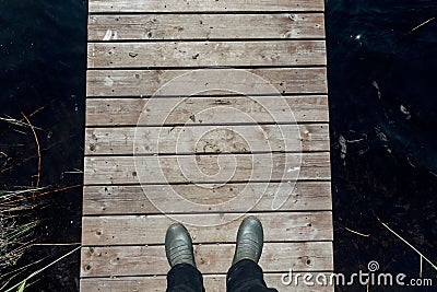 Top view of a person wearing green rubber boots standing on a wooden deck on water Stock Photo