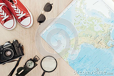 Top view of red gumshoes, film camera, magnifier, compass, sunglasses and world map on wooden surface. Stock Photo