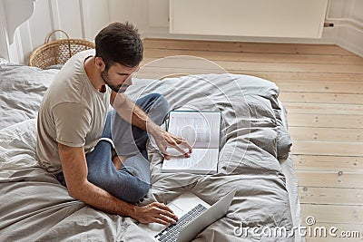 Top view of relaxed bearded guy poses at cozy bed in lotus pose, ponders on readed material, checks information from Stock Photo